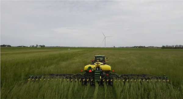 A green tractor planting in a no-till field with a wind turbine in the distance