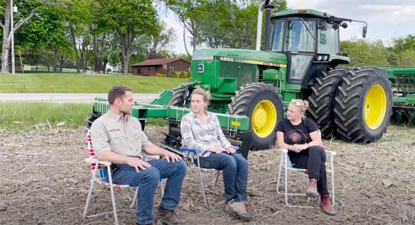 Man and two women sitting in lawn chairs in front of farm equipment