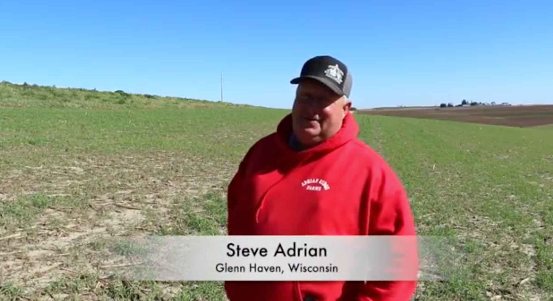 A man in a red sweatshirt talking to the camera from a farm field